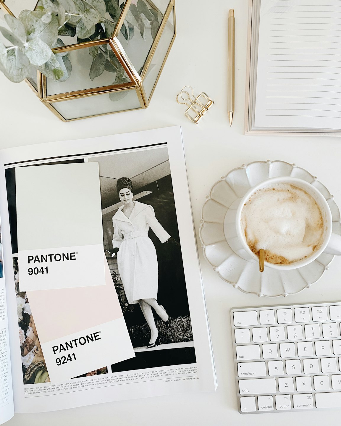 Scrivania bianca con rivista aperta, caffè, tastiera e penna dorata.