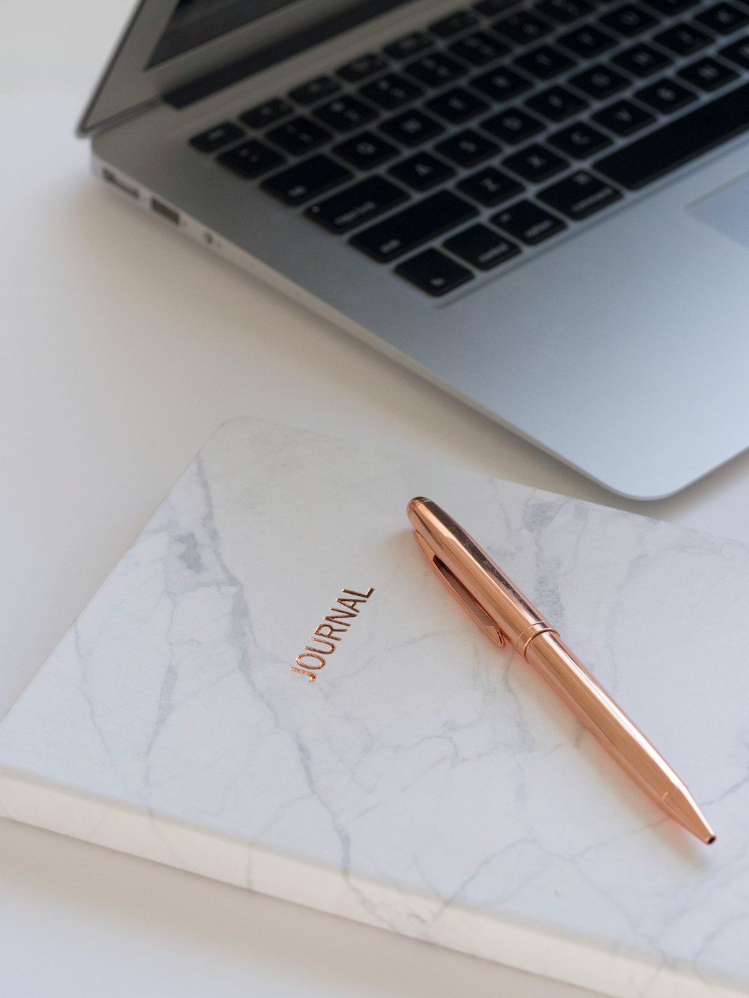 Scrivania minimalista con laptop argentato, diario color oro rosa e penna.