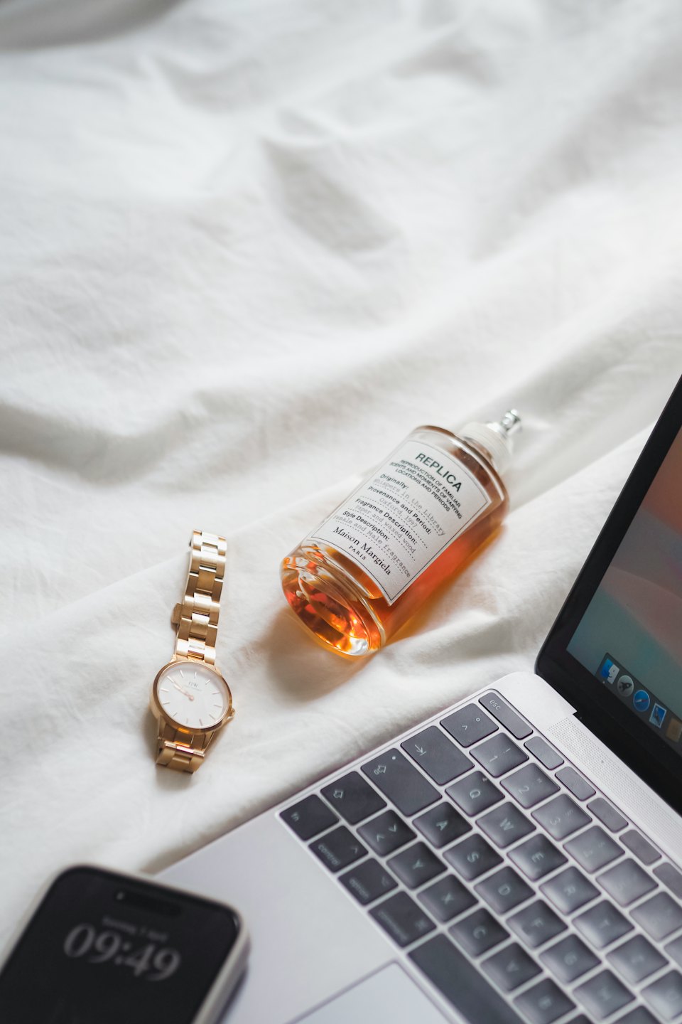 Un laptop, un orologio da polso, uno smartphone e un profumo su lenzuola bianche.